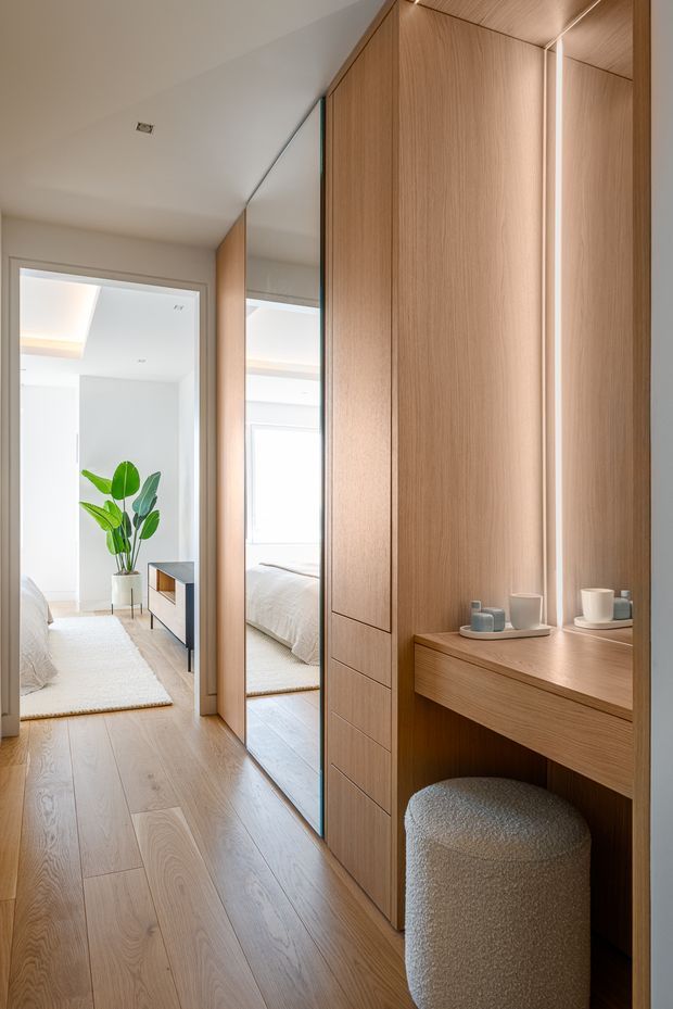 A bright, refined bedroom dressing area with full-height light timber built-in cabinetry, mirrored wardrobe doors, and an integrated timber vanity desk. A softly upholstered rounded stool sits beneath the desk, while the mirror reflects the adjacent bedroom with white bedding, pale walls, and a potted plant. Light timber flooring and warm concealed lighting create a calm, cohesive interior.