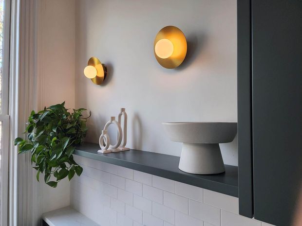 A refined bathroom vanity and display niche with a dark stone or composite countertop, white subway-tile wall finish, sculptural white vessel basin, and dark built-in cabinetry. Warm brass-gold wall sconces, decorative ceramics, and a leafy plant add contrast. Soft daylight enters from the left, creating a calm, contemporary atmosphere.