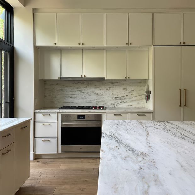 Bright, refined kitchen with cream-white Shaker-style cabinetry, tall integrated storage, stainless-steel oven and cooktop, and a large marble island. The marble backsplash continues across the cooking wall, while warm timber flooring and black-framed glazing add contrast.