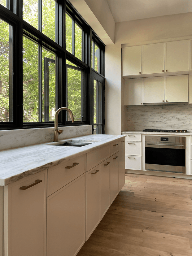 Bright, refined kitchen featuring a large central island with a pale stone benchtop, integrated sink and brushed brass-toned tapware. White built-in cabinetry lines the rear wall around a stainless-steel oven and cooktop, with a pale stone or marble-look splashback. Full-height black-framed windows bring in abundant natural light and overlook leafy trees. The room has warm timber flooring and a clean, understated contemporary character.