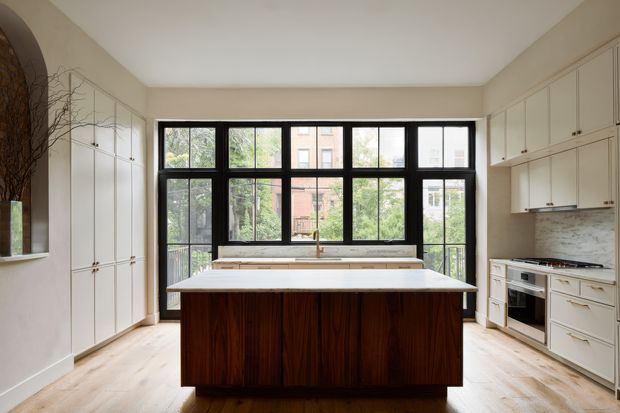 Bright, refined kitchen with a large dark timber island centred in the room, pale bespoke cabinetry along both side walls, light timber flooring, and a broad black-framed multi-pane window wall overlooking greenery and neighbouring buildings. The kitchen includes integrated appliances, a marble-look or stone splashback, and a simple contemporary fit-out with traditional detailing in the cabinetry.