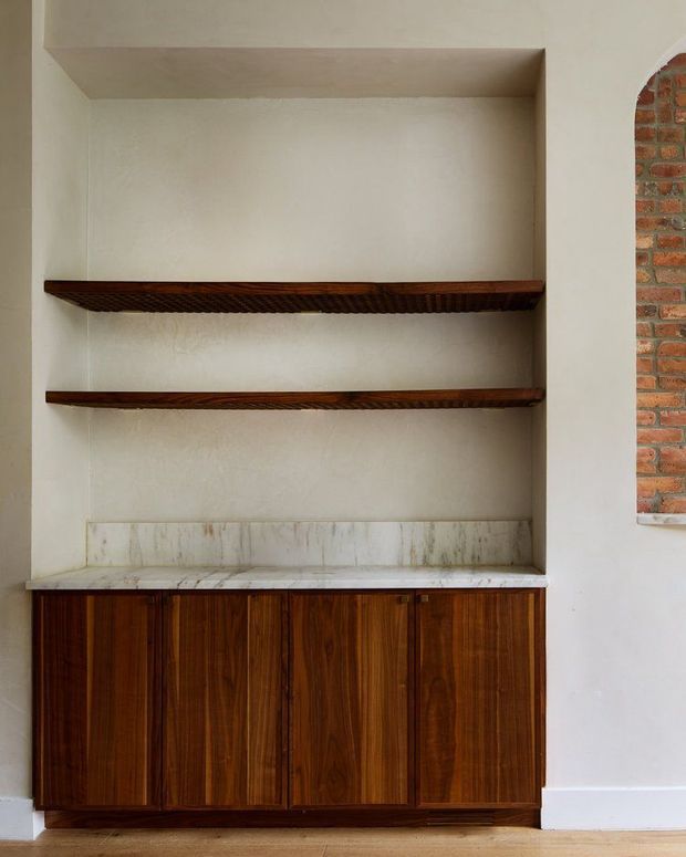 A built-in wall niche with two dark timber display shelves above a marble or stone countertop and backsplash, supported by rich dark-wood cabinetry with vertical panelled doors. The surrounding walls are light textured plaster, and an exposed red-brick arched opening is visible at the right edge. The image focuses on custom joinery and shelving in a warm, refined interior.