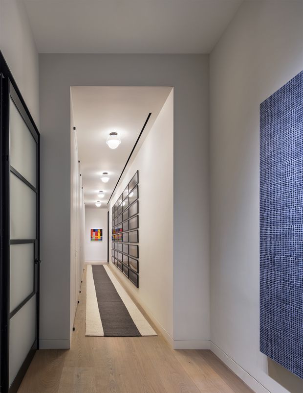 A narrow, contemporary residential hallway viewed from an adjoining space. The corridor has pale timber flooring, smooth light-grey walls, a white ceiling with a row of round flush-mounted lights, black-framed glazed doors on the left, and a large black-framed glass artwork or display installation along the right wall. A blue mosaic-tile feature surface is visible in the foreground on the right, while colourful artwork accents the far end. A long neutral runner emphasises the hallway perspective.
