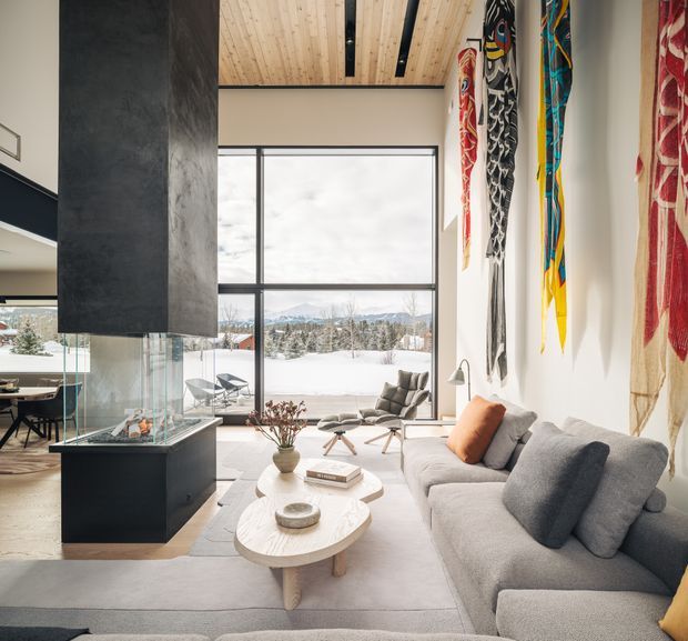 A bright contemporary living room with a large grey sectional sofa, sculptural light-wood coffee tables, lounge chairs, and a suspended dark fireplace surround. Floor-to-ceiling glazing frames a snowy landscape, while colourful long-form wall textiles provide bold vertical accents. The room features a light timber ceiling with dark recessed beams and an open connection to a dining area.