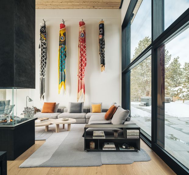 Contemporary living room with a large grey sectional sofa, layered round timber coffee tables, a black open shelving console, and colourful elongated wall sculptures. Full-height black-framed glazing opens the room visually to a snowy wooded exterior, while a warm timber-lined ceiling and pale timber floor add natural contrast.