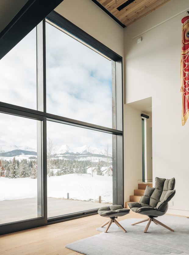 A bright, refined living room with expansive floor-to-ceiling black-framed glazing overlooking a snowy mountain landscape. The room features pale timber flooring, a warm timber-lined sloped ceiling, light walls, and a sculptural upholstered lounge chair with matching ottoman. A colourful textile hangs near a recessed doorway, adding a small decorative accent.