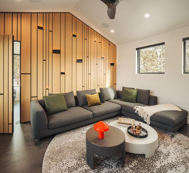 A contemporary living room with a large charcoal L-shaped sectional sofa, layered green and mustard cushions, round nesting coffee tables, and a patterned area rug. The room features a dramatic full-height timber wall with narrow vertical black reveals and integrated concealed doors, white walls, recessed ceiling lights, a ceiling fan, and dark flooring. Black-framed windows bring in natural light and views of trees.