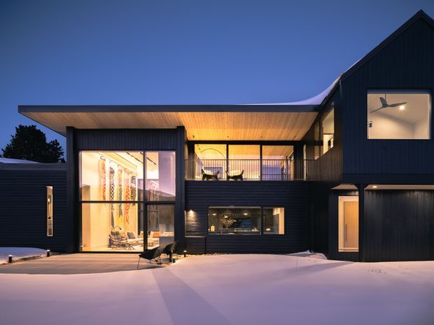 Dusk exterior view of a contemporary multi-level house in a snowy setting. The dark vertical-clad facade combines a steep gabled upper volume with a broad low-slung roof and deep timber-lined eaves. Large areas of glazing reveal warmly lit interiors, including an upper-level balcony or living area and a tall glazed space, while snow covers the foreground.