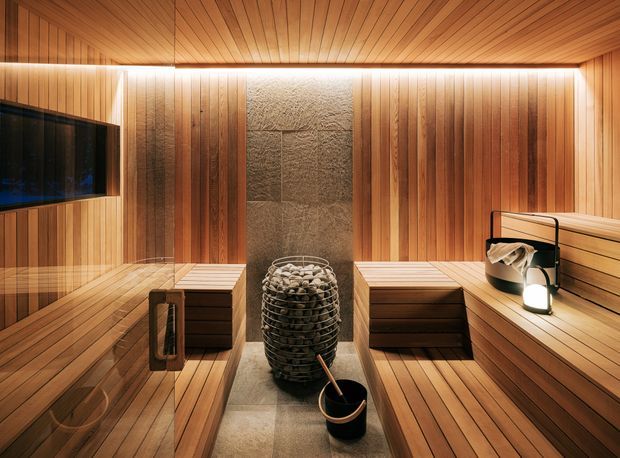 A warmly lit, compact sauna clad in vertical and horizontal timber panelling, with tiered wooden benches along the sides, a central stone-filled sauna heater, and a bucket and ladle in the foreground. A dark glass screen or television is visible on the left wall, while concealed linear lighting highlights the timber ceiling and walls.