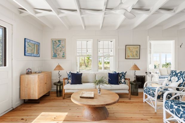 Bright, inviting coastal living room with white walls, exposed white-painted ceiling beams, timber plank flooring, and abundant natural light. The room includes a cream sofa, blue-and-white patterned armchairs, a round timber coffee table, a timber sideboard, lamps, framed artwork, and a ceiling fan. Windows and a glazed door provide views to greenery and water beyond.