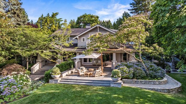 A landscaped exterior view of a large, traditional timber-clad suburban house set within a mature garden. The home has a pitched gabled roof, covered veranda, multiple levels, timber-framed windows, broad steps, and a raised stone-edged patio with outdoor seating. Dense planting, flowering shrubs, lawn, and established trees surround the house.