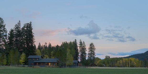 A long, low dark-clad rural house or lodge sits beyond a broad green field, with a pitched roof, illuminated windows, fencing, and dense forested hills behind it under a blue-pink evening sky.