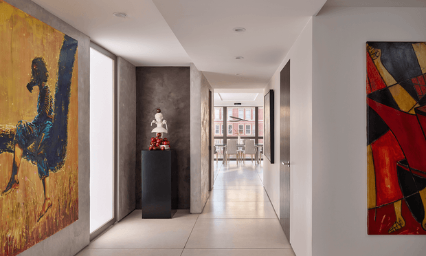 A sleek contemporary interior hallway with a long, bright linear layout, pale large-format floor tiles, smooth white walls, recessed ceiling downlights, and full-height glazing along the left side. Abstract artworks frame the corridor, while a dark textured feature niche contains sculptural decor. The hallway leads toward a dining area visible at the far end through glass doors or glazing.