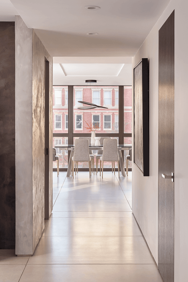 A minimalist interior view down a hallway into a dining area. The corridor has smooth pale walls, large pale floor tiles, recessed ceiling lights, and a dark wood-toned wall or panel on the left. An open door with a slim metal handle and a large dark-framed artwork or panel leads toward a bright dining space. The dining area features a simple table, upholstered chairs, a linear pendant light, and full-height windows overlooking a historic brick building. A recessed ceiling lightwell adds architectural interest.