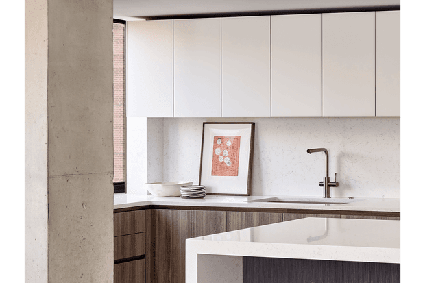 A refined contemporary kitchen viewed past a raw concrete structural column. The space features a white stone island with a waterfall edge, dark vertically grained timber cabinetry, white full-height overhead cabinets, a pale textured splashback, and a dark metal tap. A narrow window opening and framed artwork add depth, while the restrained palette and clean lines create a calm, minimalist feel.