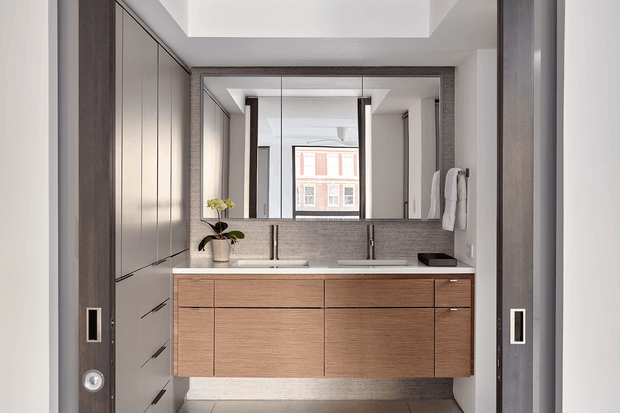 Contemporary bathroom vanity area with a wide double-sink timber vanity, dark stone-look benchtop and matching framed mirror. Full-height dark cabinetry and dark trim frame the space, while wall-mounted towels, a small orchid, recessed ceiling detailing and a window reflected in the mirror add practical and decorative elements. The room is bright, refined and highly tailored.