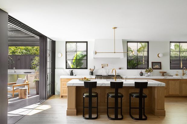 Bright, refined contemporary kitchen with a large central island and three black bar stools. The room features warm timber cabinetry, white marble-look benchtops and splashback, black-framed windows, brass tapware and pendant lighting, and expansive folding glass doors opening to a covered outdoor area.