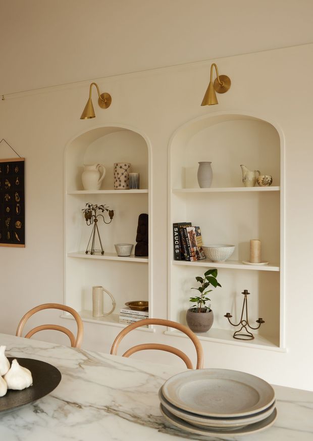 Warm, refined dining nook with a white marble dining table in the foreground, curved timber dining chairs, and two arched built-in display niches set into a cream wall. The shelves hold ceramics, books, decorative objects, candles, and a small potted plant, while brass wall sconces provide soft accent lighting.