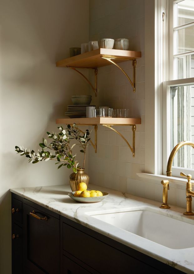 A warm, refined kitchen vignette featuring a deep white farmhouse-style sink beneath a tall multi-pane window, dark painted cabinetry with brass hardware, and a white marble countertop. Two open timber shelves with ornate brass brackets display ceramics and glassware against a light tiled backsplash. A brass faucet, a bowl of lemons, and a leafy branch add classic decorative detail.