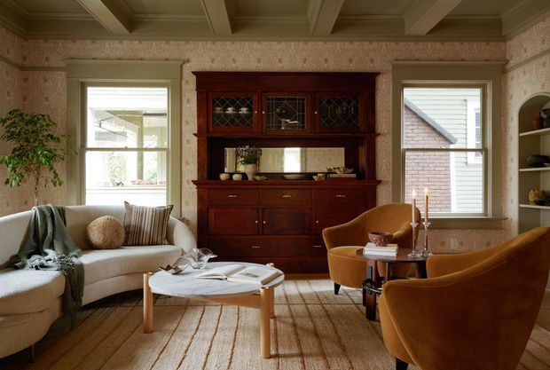 Warm, traditionally styled living room with a cream sofa, two ochre velvet armchairs, a light timber coffee table, and a large dark-stained built-in hutch or cabinetry unit. The room features patterned wallpaper, timber flooring, a coffered timber ceiling, white-trimmed windows, open shelving, and layered neutral textiles. Natural light enters through the windows, creating a refined and inviting atmosphere.