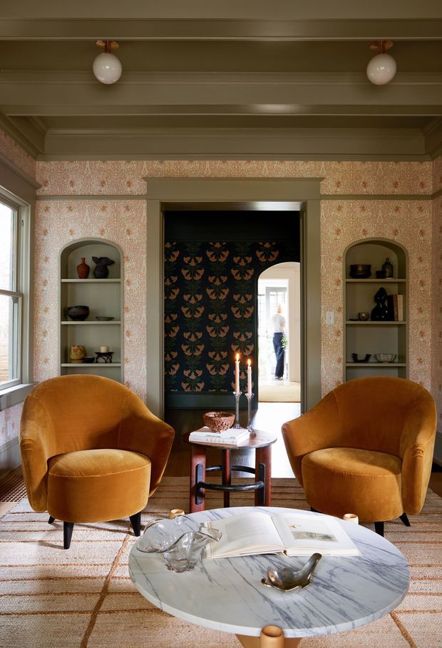 A richly styled sitting room with two curved mustard velvet armchairs facing a round marble coffee table. The room features ornate patterned wallpaper, built-in arched display niches, dark floral wallpaper in the doorway, decorative wall and ceiling mouldings, vintage-inspired pendant lights, and layered neutral carpeting. A hallway and adjoining bright room are visible beyond the central doorway.