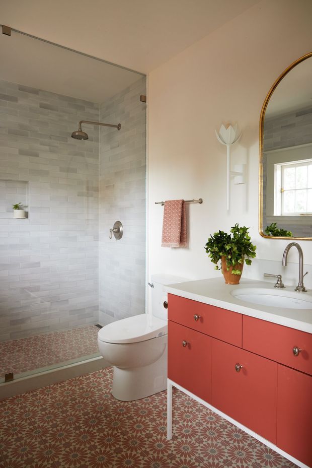A bright bathroom with a glass-enclosed walk-in shower, pale grey rectangular wall tiles, a patterned terracotta and cream floor, a white toilet, and a coral-red vanity with a white countertop and integrated basin. A brass-framed mirror, brass tapware, a towel rail, wall sconce, and potted plant add warm decorative detail.