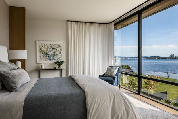 A refined contemporary bedroom with a large upholstered bed, layered grey and white bedding, a blue-grey accent chair, and a slim console beneath abstract artwork. Full-height curtains and expansive corner glazing overlook a waterfront, with warm timber wall panelling and flooring adding contrast to the light neutral interior.