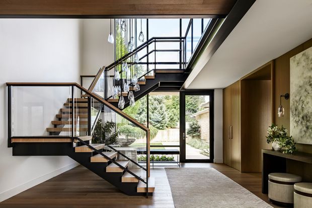 A refined modern interior foyer with a dramatic open staircase, black metal framing, timber treads, and clear glass balustrades. The space features warm timber ceiling and flooring, a large neutral rug, full-height glazing and a glazed door opening to a landscaped courtyard. Built-in timber wall joinery, pendant lights, a console, artwork, and stools complete the sophisticated entry space.