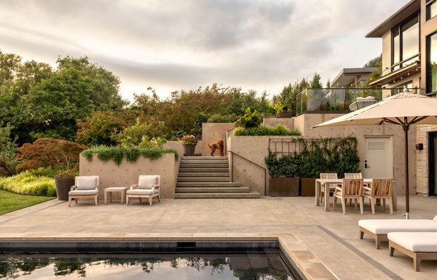 A landscaped contemporary residential courtyard with a large paved terrace, swimming pool, concrete retaining walls and steps, outdoor dining furniture, lounge chairs, umbrellas, and dense garden planting. A multi-level modern home with extensive glazing is visible at right.