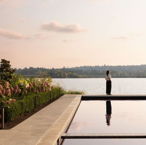 A landscaped waterfront terrace with a long rectangular reflecting or swimming pool in the foreground, bordered by a broad concrete-paved walkway and low dark metal handrails. Dense flowering planting lines the left edge, while a person stands on the far pool edge overlooking the calm lake and wooded opposite shore. The image has soft warm evening light and a tranquil, refined outdoor atmosphere.