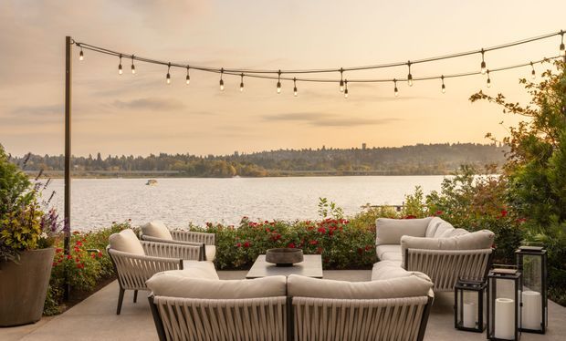 A stylish waterfront outdoor lounge terrace overlooking a calm lake, furnished with cream rope-woven sofas and armchairs around a low coffee table. String lights are suspended overhead, with planters, flowering shrubs, and lanterns creating a warm evening entertaining setting. A wooded shoreline is visible across the water.