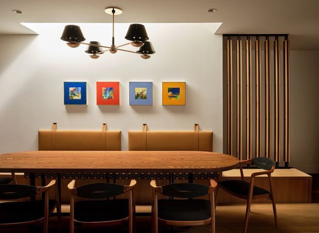 Warm, mid-century-inspired dining room with a long solid timber dining table, black upholstered wooden chairs, built-in tan leather banquette seating, colourful framed artwork, a sculptural multi-arm pendant light, and a vertical timber slatted screen on the right. A large ceiling skylight or recessed light well brightens the otherwise intimate space.