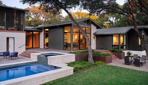 Exterior view of a contemporary, single-storey residence arranged around a landscaped outdoor entertaining area. The home features dark vertical cladding, low-pitched metal roofs, extensive floor-to-ceiling glazing, warm interior lighting, pale masonry walls, timber decking, a swimming pool and raised spa, with mature trees integrated into the garden.
