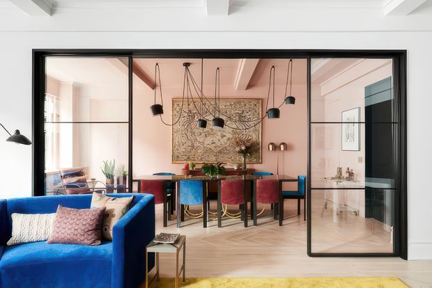 A refined open-plan interior viewed from a living room, with a vivid blue sofa in the foreground and a dining area separated by black-framed glazed partition doors. The dining space has a dark wood table, colourful upholstered chairs, sculptural pendant lighting, patterned wall art, and warm pink walls. Pale herringbone timber flooring, white exposed ceiling beams, and a glazed opening toward a compact bathroom or washroom add architectural interest.