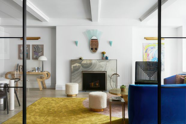 Bright, eclectic contemporary living room viewed through black-framed internal glazing. The room features a sculptural fireplace surround, white walls and ceiling with exposed structural beams, vivid blue seating, a mustard area rug, a glass-topped coffee table, and distinctive mid-century and art-inspired furnishings. A small dining or work area is visible to the left.