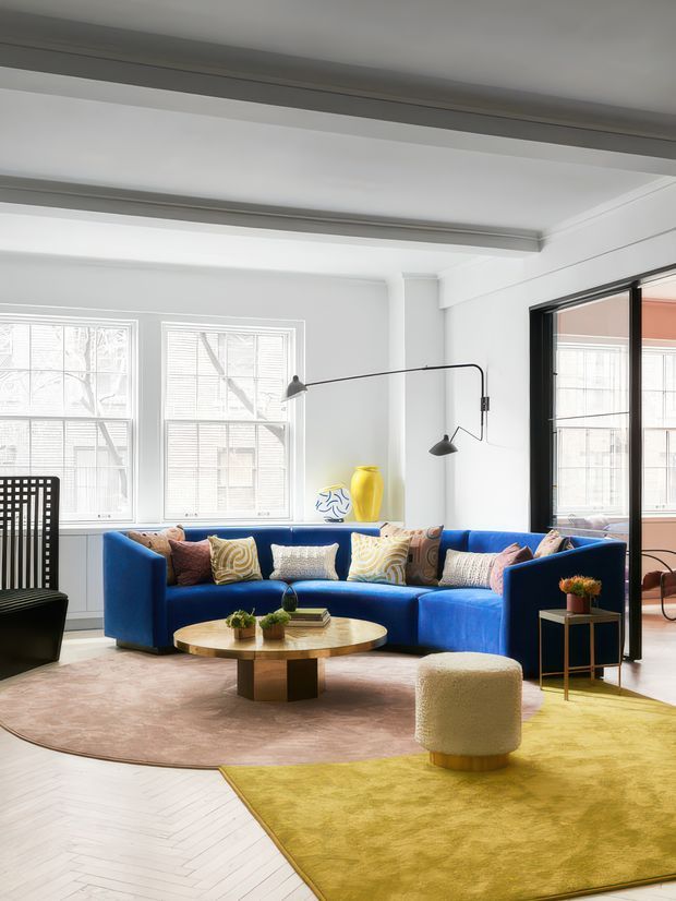 Bright, contemporary living room with white walls, exposed ceiling beams, tall multi-pane windows, and black-framed glazed doors. A vivid cobalt-blue sectional sofa is arranged around a round wood coffee table, with colourful cushions, sculptural decor, a black lounge chair, side table, floor lamps, and layered blush and mustard area rugs. The room has an eclectic, artful and inviting feel.