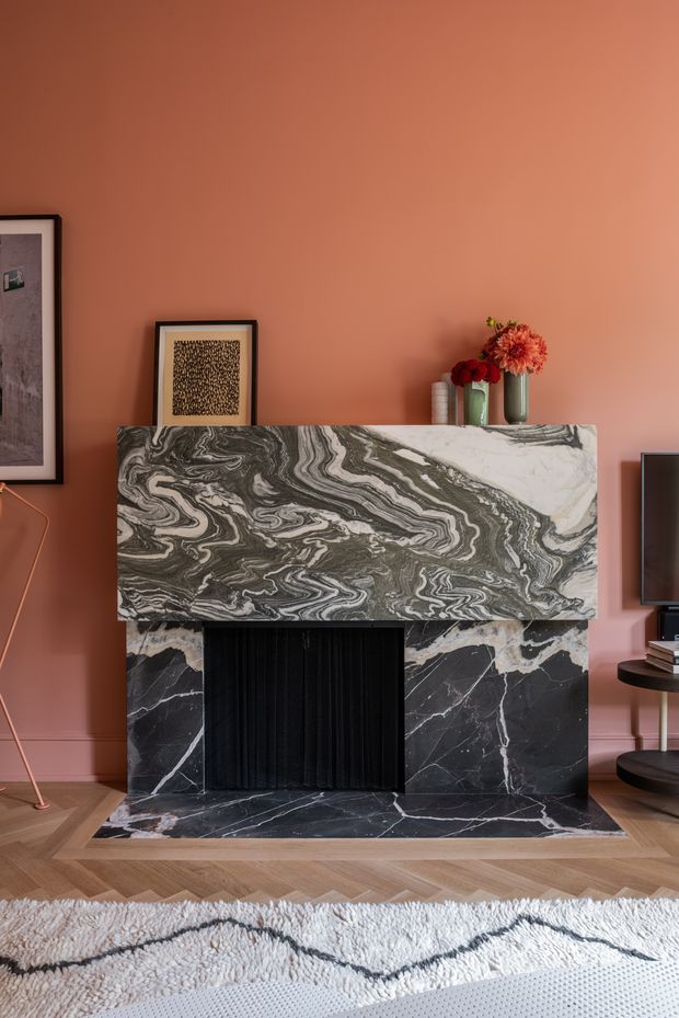 A close-up interior view centred on an ornate fireplace with dramatic black-and-white veined marble cladding and a heavily patterned marble mantel. The fireplace is set against a warm terracotta-pink wall, with herringbone timber flooring, a textured rug, framed artwork, flowers, and a small television console visible at the sides. The room has a bold, refined, decorative character.