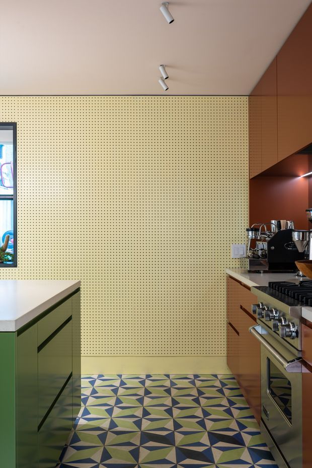 A narrow, contemporary kitchen viewed toward a perforated cream feature wall. The room has green lower cabinetry with dark linear pulls on the left, warm timber-toned cabinetry on the right, a stainless-steel range and commercial-style coffee machine, white benchtops, exposed directional spotlights, and a bold geometric patterned tile floor in blue, green, cream, and dark tones.