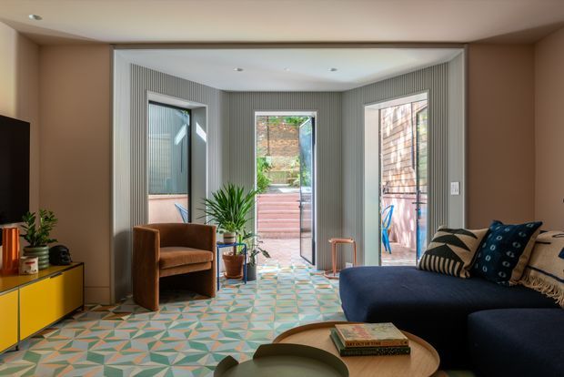 A bright, colourful living room with a deep navy sectional sofa, brown armchair, yellow low media cabinet, indoor plants, and a sculptural wood coffee table. The room features a geometric multicoloured tiled floor, vertically ribbed grey wall panelling, recessed ceiling lights, and large glazed doors and windows opening toward an outdoor deck or courtyard.