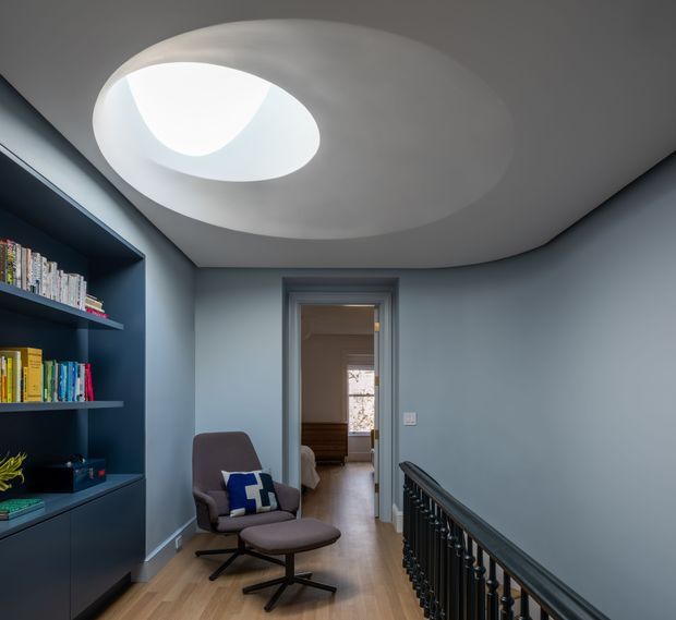 A refined upstairs landing or hallway with a curved sculptural ceiling and large circular skylight. The space features blue-grey painted walls, built-in dark shelving filled with books, a lounge chair and ottoman, light timber flooring, and a dark decorative stair balustrade. A bedroom is visible through the doorway at the end of the hall.