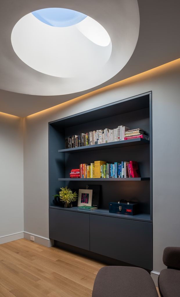 A refined modern interior featuring a dark built-in bookcase with open shelving, lower cabinetry, books, framed artwork and a small plant. The joinery is set into a light grey wall and paired with warm concealed LED strip lighting, a sculptural circular ceiling opening or skylight, and light timber flooring. Part of a dark upholstered seating area is visible in the foreground.