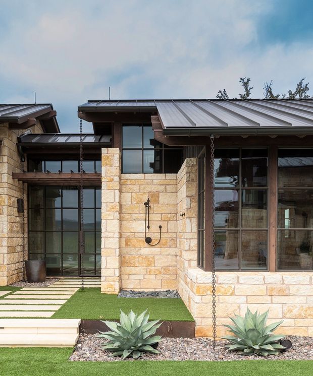 Exterior view of a low-slung contemporary building composed of warm pale stone masonry, dark timber-framed glazing, and broad standing-seam metal roofs. The landscaped foreground includes clipped lawn, gravel beds, agave plants, stepping stones, and exposed rain chains. Large glazed openings reveal interior spaces but the building facade and courtyard landscaping dominate the image.