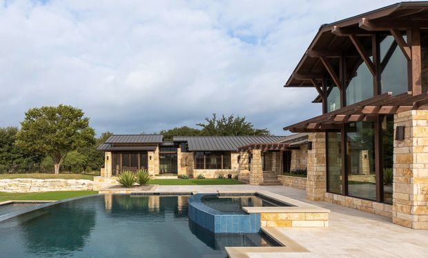 Exterior view of a sprawling low-rise stone-clad residence arranged around a swimming pool and raised spa. The building features dark metal pitched roofs, extensive glazing, exposed timber post-and-beam framing, deep eaves, and covered outdoor areas. A paved pool terrace and landscaped grounds are visible in the foreground.
