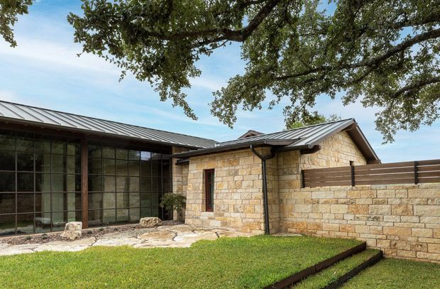 Single-storey stone-clad building with a low-pitched metal roof, large dark-framed glazed wall, and a small grass courtyard. A low stone privacy wall and horizontal timber screening extend from the building, while mature tree branches frame the upper edge of the image. The architecture has a restrained contemporary character with strong indoor-outdoor glazing and natural masonry.