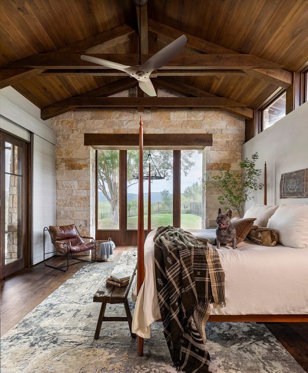 Rustic yet refined bedroom with a vaulted timber ceiling, exposed dark wood beams, a natural stone feature wall, and expansive timber-framed glazing overlooking greenery. The room includes a large upholstered bed, leather lounge chair, patterned area rug, wood coffee table, ceiling fan, and warm earthy furnishings.