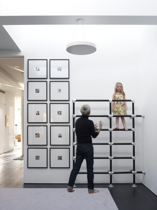 Bright contemporary interior featuring a tall white wall with a grid of black-framed artworks, a large circular pendant light, and a black modular climbing/play structure being used by an adult and child. An adjacent hallway and living area are visible to the left. The space has pale walls, dark timber flooring, and a large light rug.