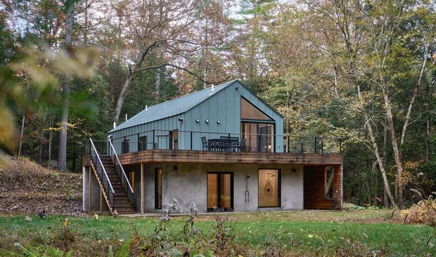 A modern two-level cabin-style house set within a wooded landscape. The building has a steep gabled metal roof, blue-grey exterior cladding, large glazed openings, a timber-framed elevated deck with dark metal railings, and an external stair descending to the lower level. The lower storey combines concrete-rendered walls with timber cladding and glazed doors. Mature deciduous trees surround the home.