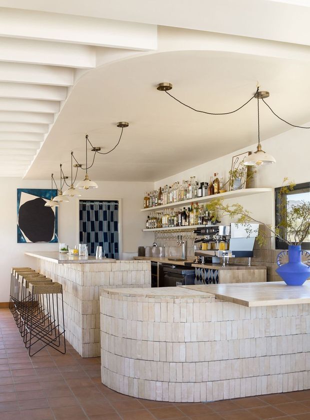 A bright, stylish bar and hospitality interior with curved bar counters clad in small pale tiles, open bottle shelving, a coffee and cocktail service area, and a row of slender black bar stools. The space features white plaster walls and ceiling, exposed stepped ceiling beams, hanging pendant lights with visible cords, terracotta-coloured floor tiles, a large mirror, patterned blue-and-white curtains, greenery, and a bold blue vase. The overall composition is warm, refined, and contemporary with strong organic curves and Mediterranean-inspired materiality.