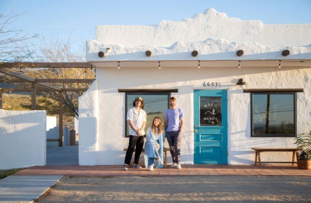A small white stucco building with a distinctive sculpted parapet roofline, dark-framed windows, and a bright turquoise entrance door. A covered pergola extends along the left side, while a brick-paved walkway and simple bench sit in front. The facade is photographed in bright sunlight against a clear blue sky, with people standing at the entrance.