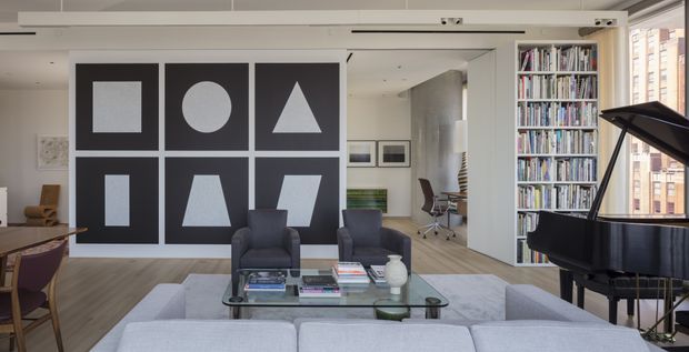 A bright, contemporary open-plan living room with a large geometric black-and-white feature wall, grey sectional sofa, two dark armchairs, glass coffee table, and a black grand piano. Floor-to-ceiling built-in bookshelves line the right side, while a compact home office is visible in the background. Pale timber flooring, white walls, recessed ceiling details, and large windows create an airy urban interior.